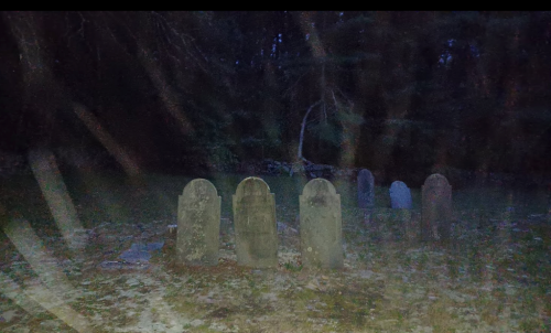 A dimly lit graveyard with several old gravestones surrounded by trees at dusk.