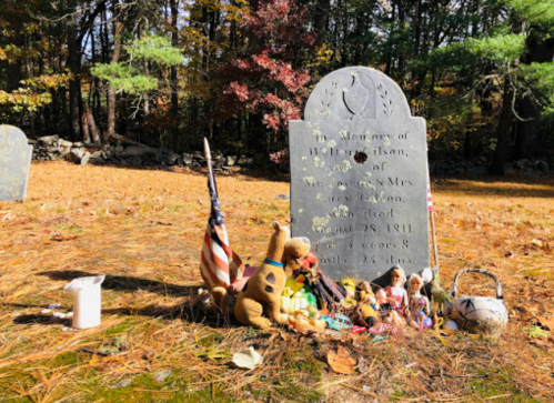 A weathered gravestone surrounded by small memorial items, including toys and an American flag, in a wooded area.