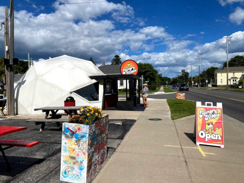 A food stand with a dome structure, picnic tables, and an "Open" sign on a sunny day with blue skies.