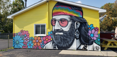 Colorful mural of a bearded man with sunglasses and a headband, surrounded by vibrant flowers on a yellow house.