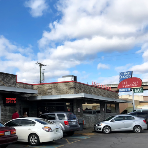 A drive-in restaurant with a retro sign, cars parked outside, and a partly cloudy sky above.