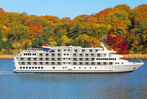 A white cruise ship glides through calm waters, surrounded by vibrant autumn foliage.
