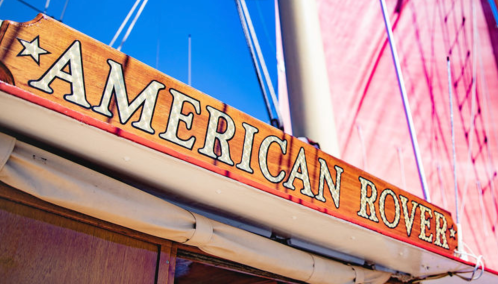 Sign reading "American Rover" on a boat, with a bright blue sky and sail in the background.