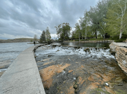 A serene riverside scene with a walkway, gentle rapids, and lush trees under a cloudy sky.