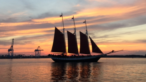 A tall ship with red sails silhouetted against a vibrant sunset over calm waters, with cranes in the background.