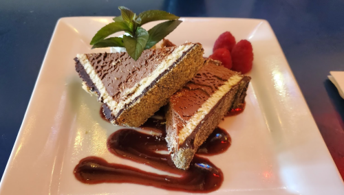 Two triangular chocolate desserts on a plate, garnished with mint leaves and raspberries, drizzled with sauce.