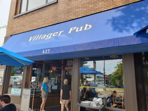 Exterior of Villager Pub with a blue awning, featuring a sign and outdoor seating area. Two people are entering.