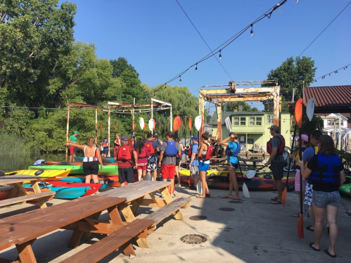 A group of people in life jackets gather near colorful kayaks by a waterfront, preparing for a kayaking activity.
