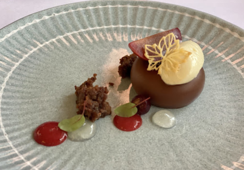 A gourmet dessert featuring a chocolate sphere, yellow mousse, and decorative sauces on a textured plate.