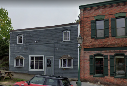 A gray wooden building next to a red brick building, with green window frames and a parked car in front.