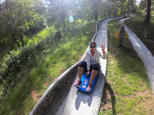 A person on a sledding track in a forest, smiling and making a peace sign, enjoying a sunny day outdoors.