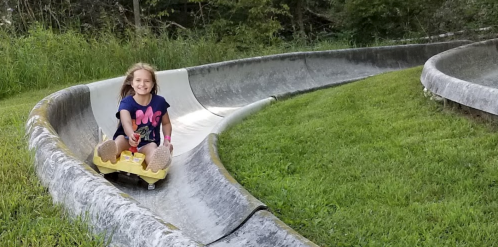 A young girl joyfully rides a yellow sled down a winding slide surrounded by green grass and trees.