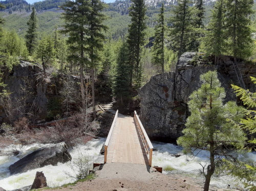 A wooden bridge spans a rushing river, surrounded by lush green trees and rocky cliffs in a mountainous landscape.