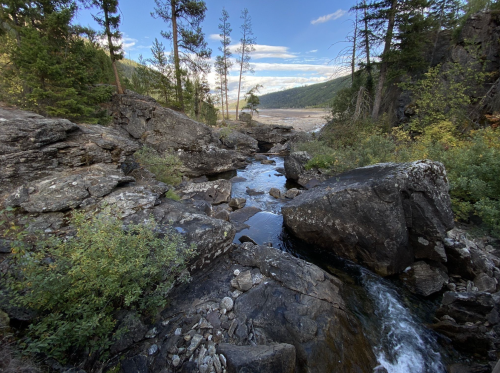 A serene landscape featuring a rocky stream surrounded by trees and mountains under a clear blue sky.