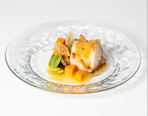 A beautifully plated dish featuring fish, colorful vegetables, and delicate garnishes on a clear glass plate.