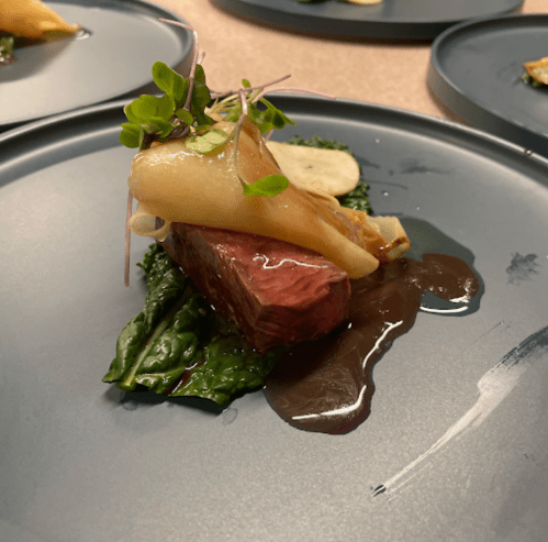 A gourmet dish featuring a piece of meat topped with vegetables and sauce, served on a dark plate.