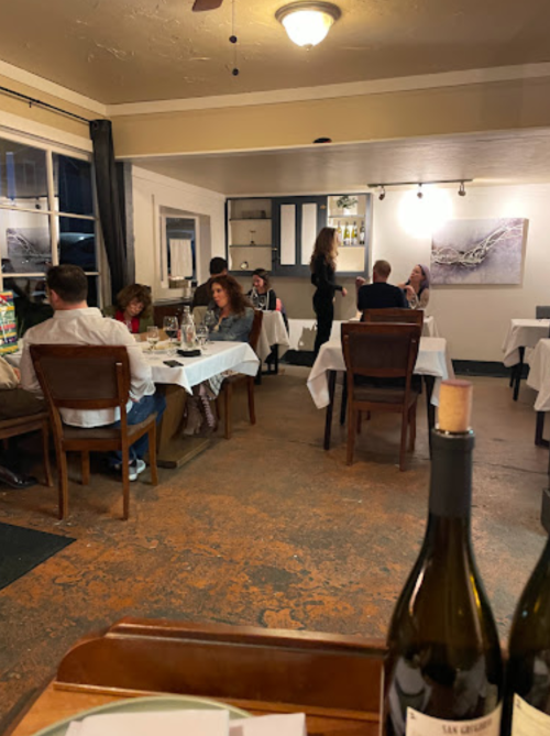 A cozy restaurant interior with diners at tables, white tablecloths, and a server attending to guests.