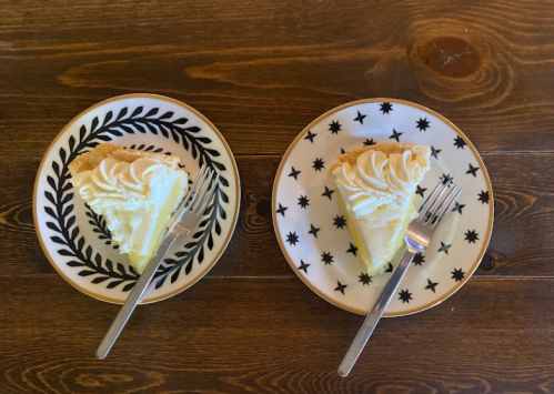 Two slices of pie with whipped cream on decorative plates, each accompanied by a fork, on a wooden table.