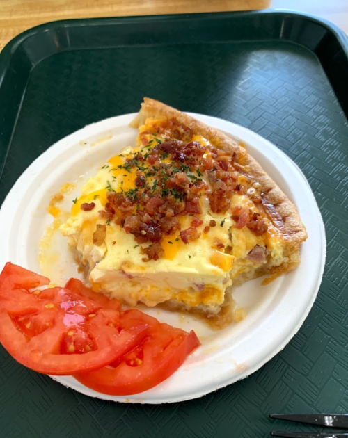 A slice of quiche topped with cheese and bacon, served on a white plate with a side of sliced tomatoes.