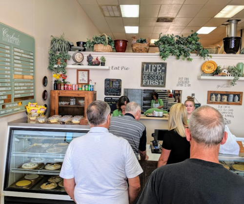 Customers line up at a cozy dessert shop, with pies displayed and a friendly staff member serving at the counter.