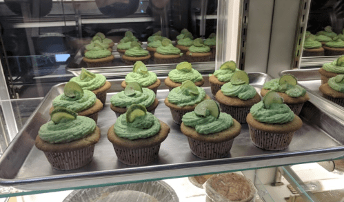 A tray of cupcakes with green frosting and lime slices, displayed in a glass case.