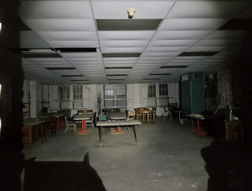 Dimly lit, abandoned room with empty tables and chairs, featuring peeling walls and a ceiling with missing tiles.