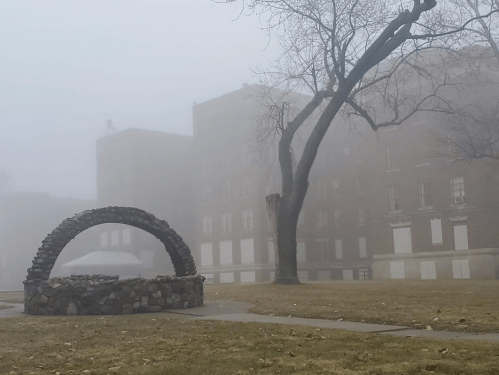 A foggy scene featuring a stone arch and a bare tree in front of a large, indistinct building.