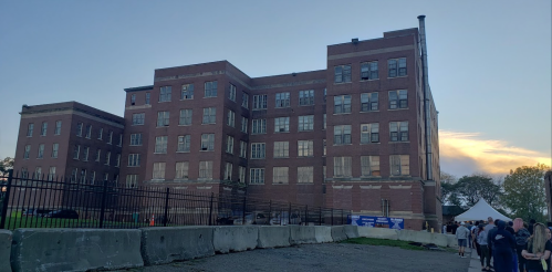 A large, old brick building with boarded windows, surrounded by a fence and a line of people in front.