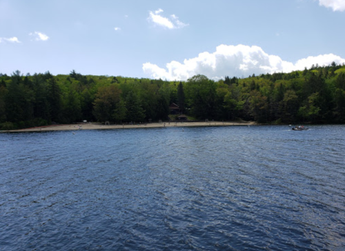 A serene lake view with a sandy beach and lush green trees under a partly cloudy sky.
