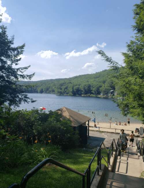 A scenic view of a lake surrounded by trees, with people enjoying the beach and a building in the foreground.