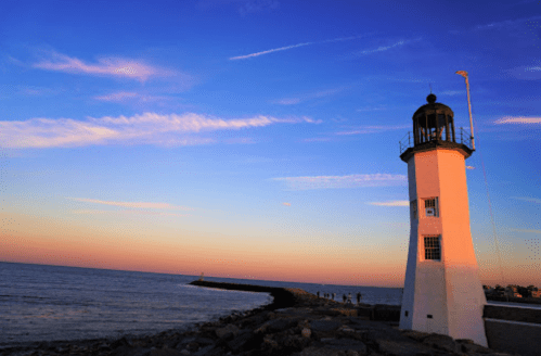 A white lighthouse stands on a rocky shore, with a colorful sunset sky and calm ocean in the background.