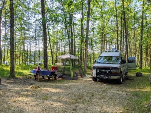 A camper van parked in a wooded area with a picnic table and a screened tent nearby, surrounded by trees.