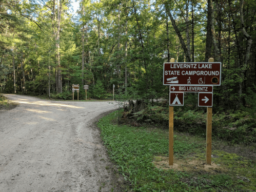 Signposts at a fork in a dirt road, directing to Leverentz Lake State Campground and Big Leverentz. Surrounded by trees.