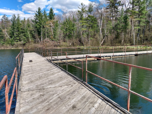 A wooden dock extends over calm water, surrounded by trees and a blue sky with clouds.