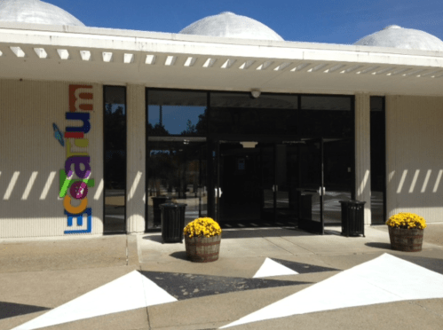 Entrance of a building with colorful signage, large glass doors, and flower pots on either side.