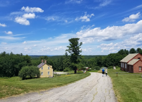 Two people walk down a gravel road with a yellow house and a red building in a scenic landscape under a blue sky.