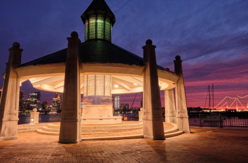 A gazebo with a lit interior against a vibrant sunset sky, overlooking a city skyline and water.