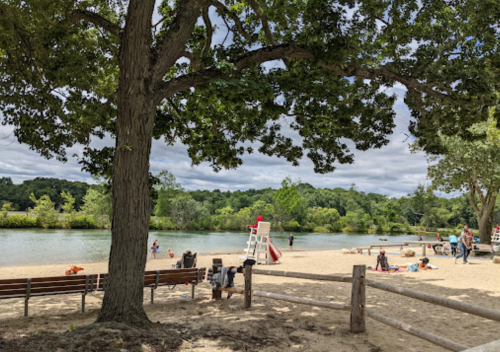 A sandy beach by a river, with people relaxing, trees, and a lifeguard stand in the background under a cloudy sky.