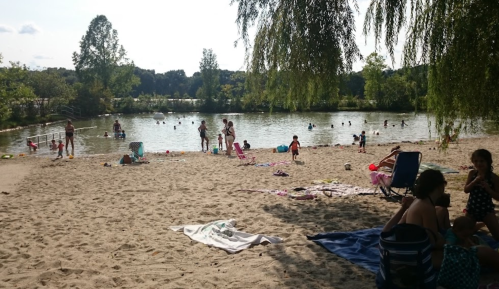 A sandy beach by a calm lake, with people swimming, sunbathing, and enjoying a sunny day outdoors.