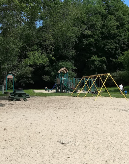 A sandy playground with swings and a climbing structure, surrounded by trees and people enjoying the space.