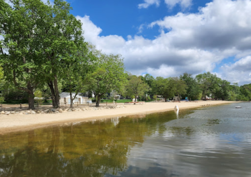 A serene beach scene with sandy shore, green trees, and calm water under a partly cloudy sky.