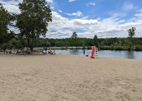 A sandy beach by a calm river, with people relaxing and a lifeguard chair in the foreground under a partly cloudy sky.