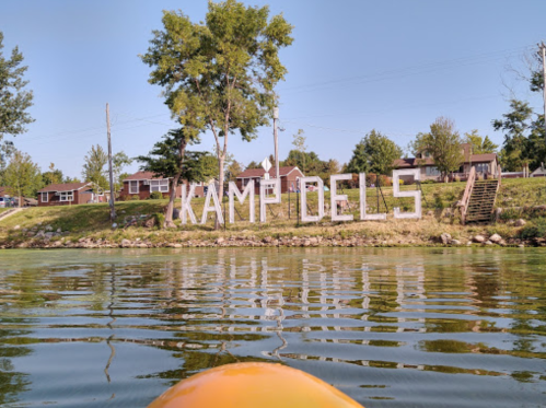View of a lake with a sign reading "Kamp Dels" on the shore, surrounded by trees and cabins in the background.