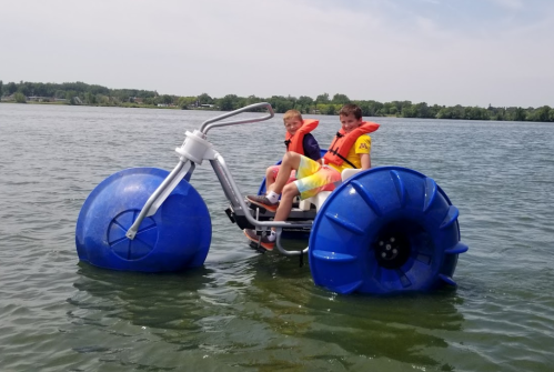 Two children in life jackets ride a water tricycle with large blue floats on a lake.