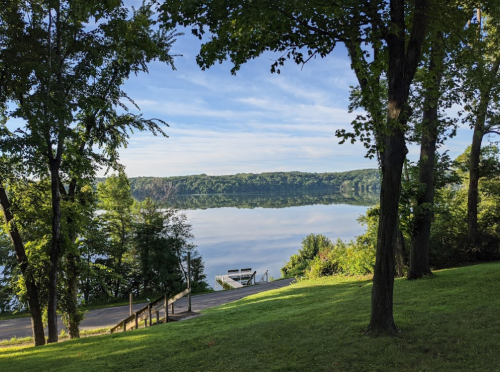 A serene lake view surrounded by trees, reflecting the sky and greenery, with a pathway leading down to the water.