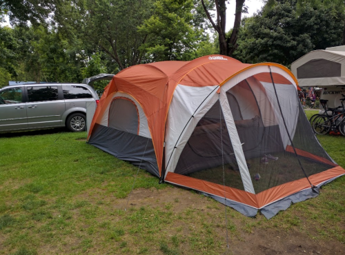 A large orange and gray tent set up on green grass, with a minivan parked nearby and trees in the background.