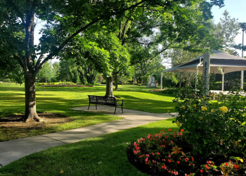 A peaceful park scene featuring a path, a bench, lush green grass, trees, and colorful flower beds.