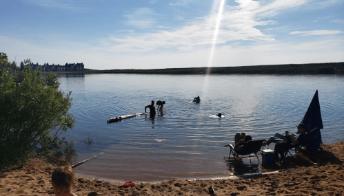 A serene beach scene with people swimming in calm water, chairs on the sand, and a clear blue sky.