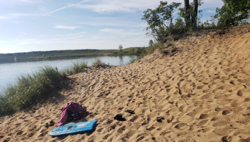 A sandy beach by a calm lake, with a backpack and a blue mat resting on the shore, surrounded by greenery.