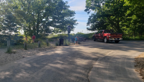 A red truck parked near a sandy path, with trees and a picnic area in the background on a sunny day.